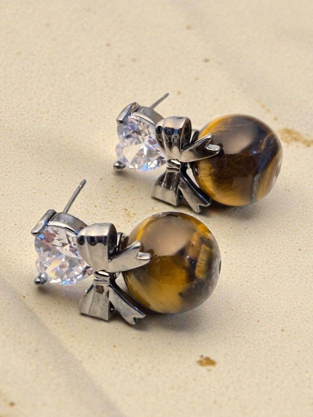Close-up of tiger eye drop stud earrings showing bow detail and golden-brown stone on sandy background