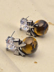 Close-up of tiger eye drop stud earrings showing bow detail and golden-brown stone on sandy background