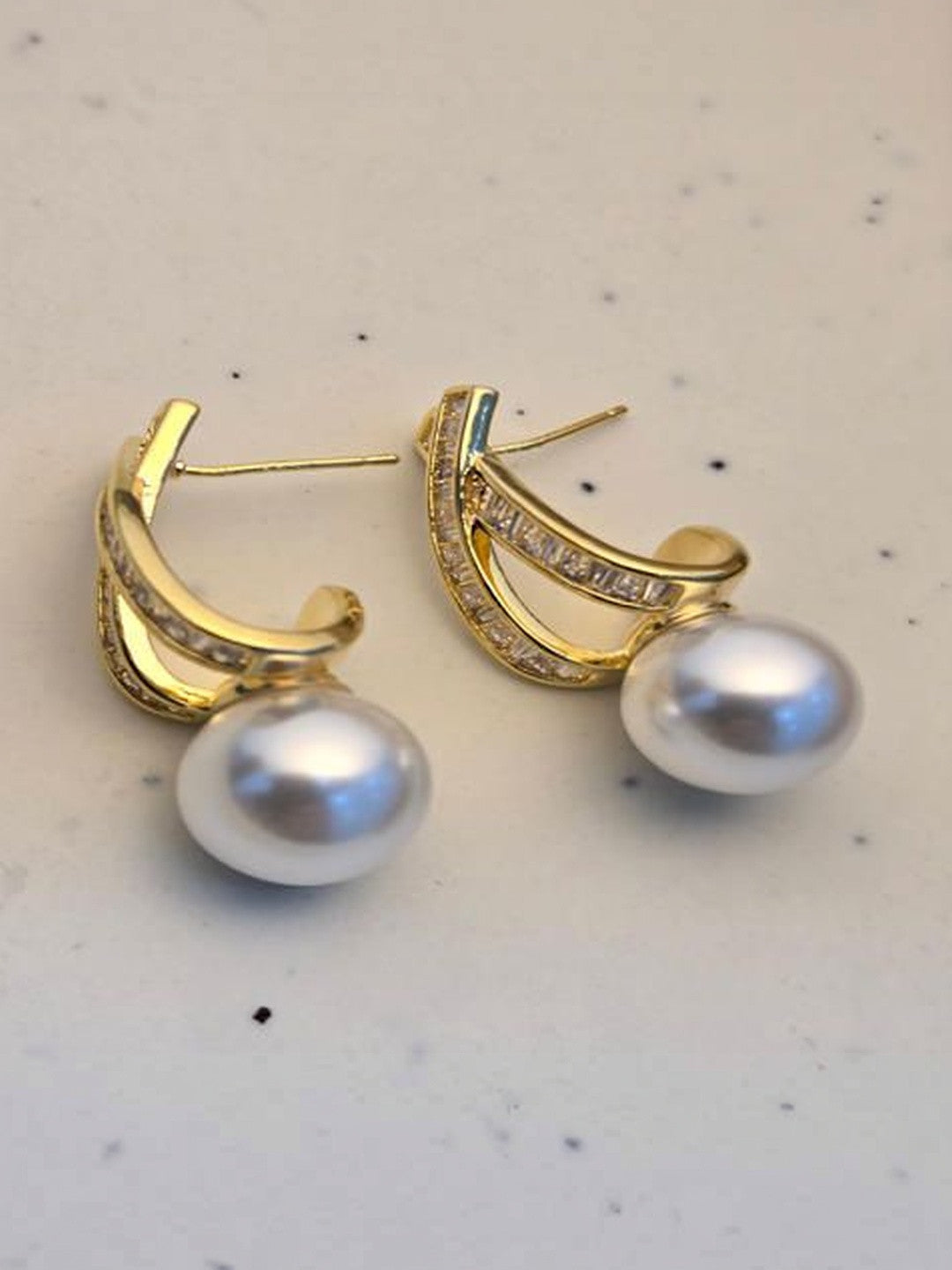 Pair of gold-plated pearl drop earrings with baguette crystals on a white marble surface, angled side view