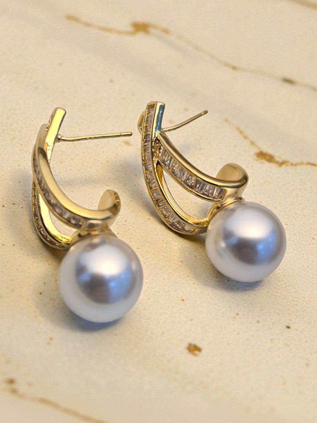 Close-up of both gold-tone pearl drop earrings resting on a warm beige marble surface showing pearl lustre