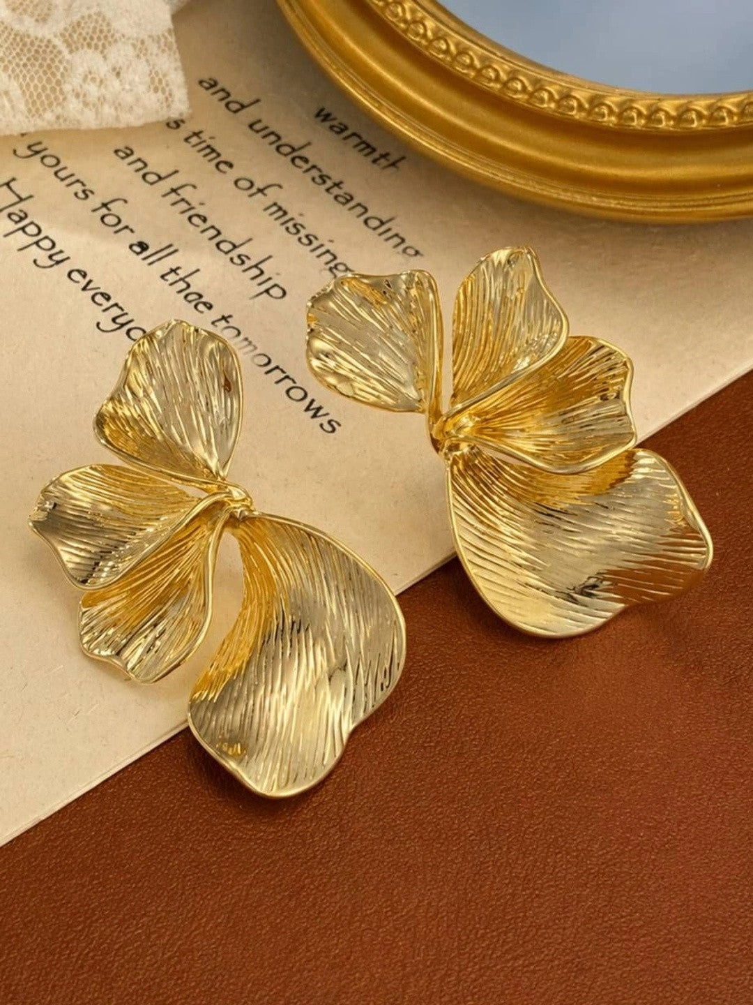 Gold-toned petal drop earring pair displayed on aged paper beside a gold-framed mirror