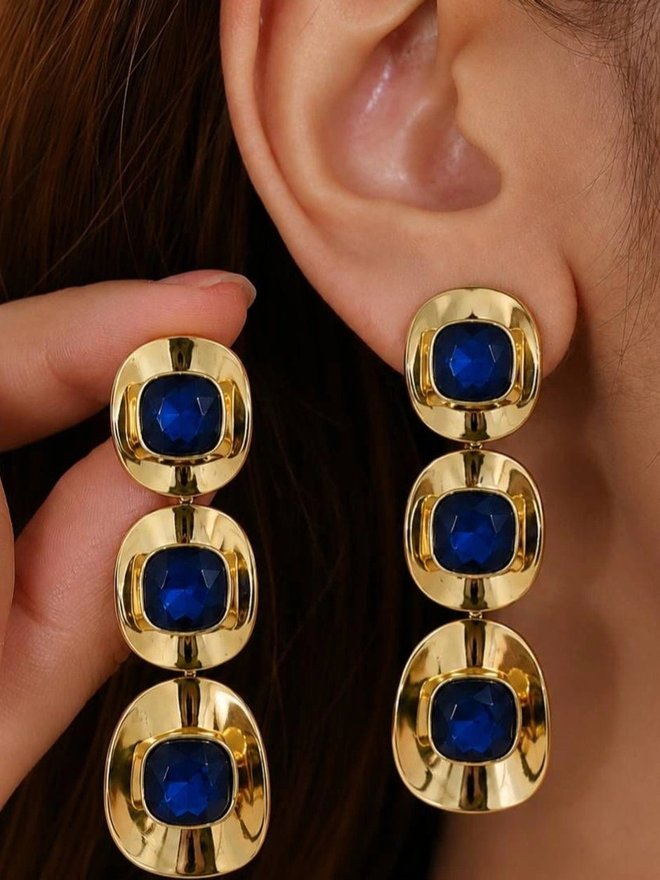 Gold-plated triple-drop blue crystal earring worn on a woman's ear, shown alongside its pair held in hand