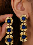 Gold-plated triple-drop blue crystal earring worn on a woman's ear, shown alongside its pair held in hand