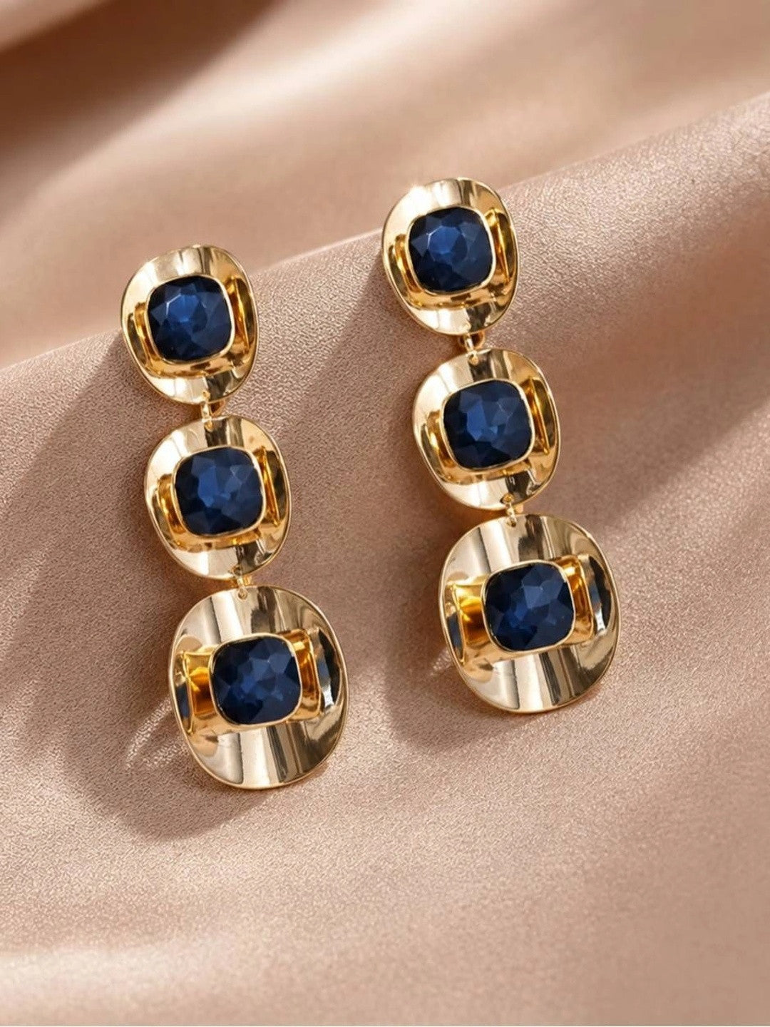 Pair of gold-plated blue crystal triple-drop earrings laid flat on rose satin fabric, front view