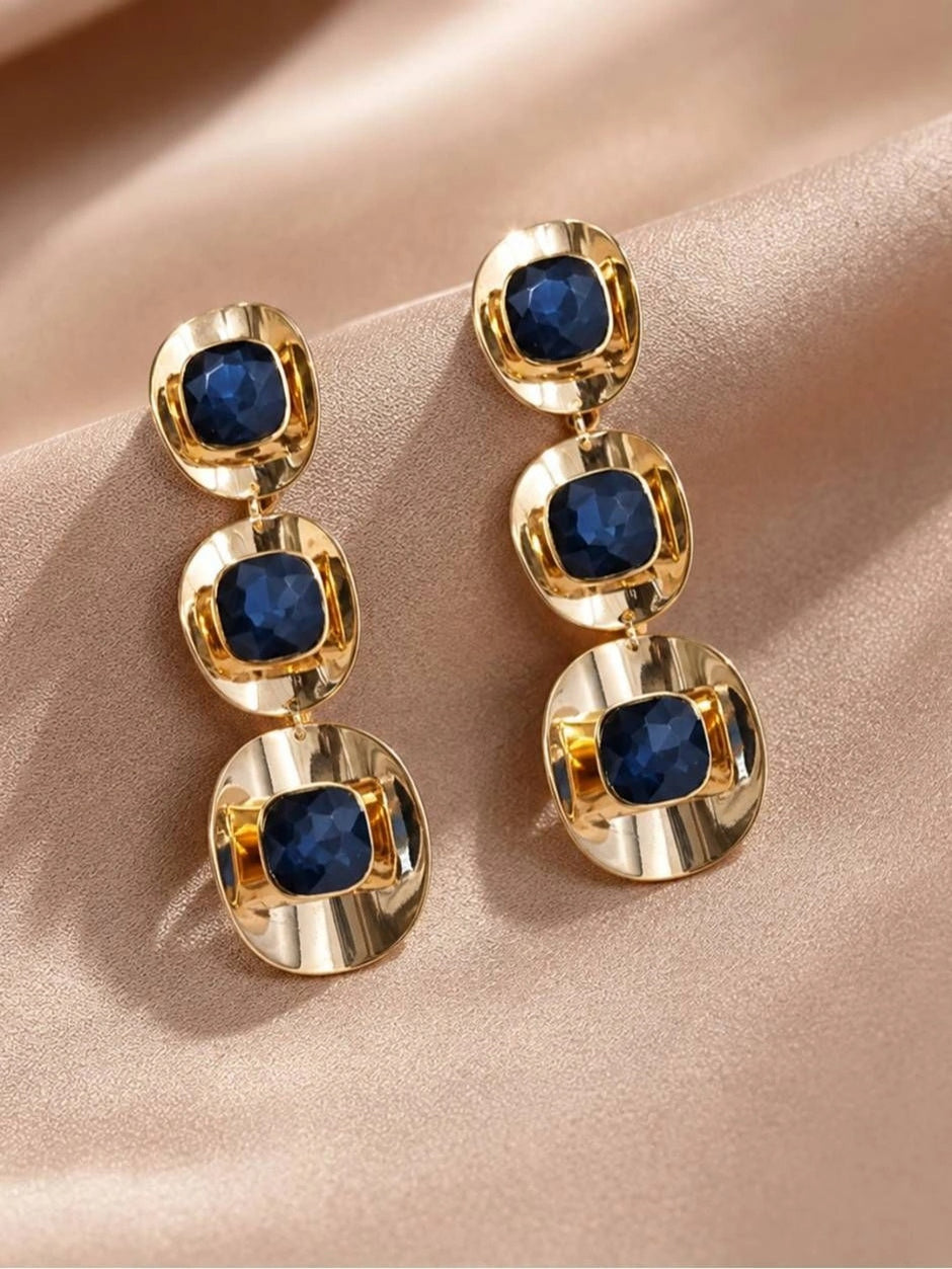 Pair of gold-plated blue crystal triple-drop earrings laid flat on rose satin fabric, front view