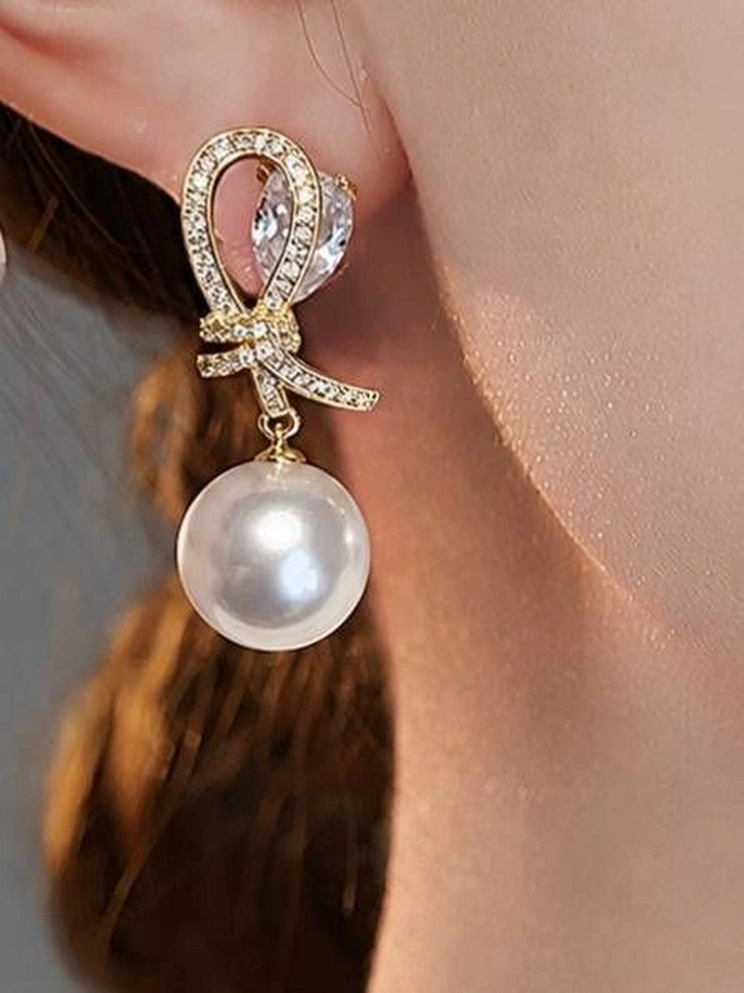 Gold-plated CZ bow and faux pearl drop earring worn on a woman's ear, side profile view