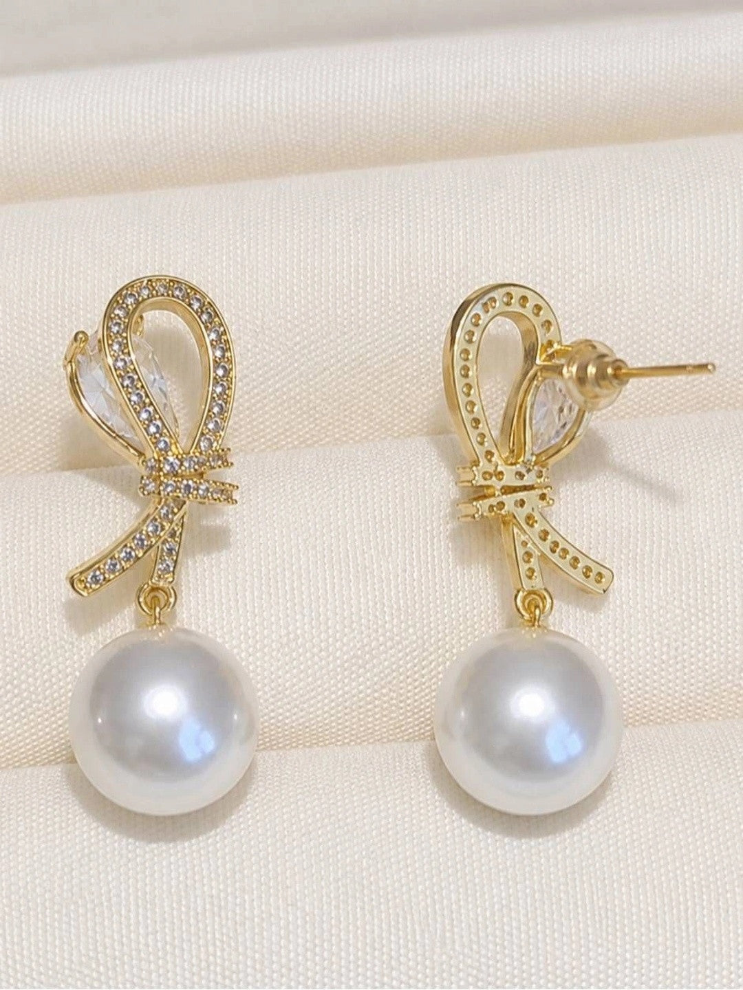 Pair of gold-plated bow earrings with pavé CZ and round pearl drops displayed on cream fabric, front view