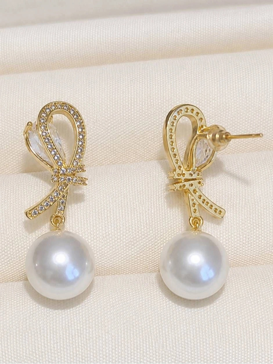 Pair of gold-plated bow earrings with pavé CZ and round pearl drops displayed on cream fabric, front view