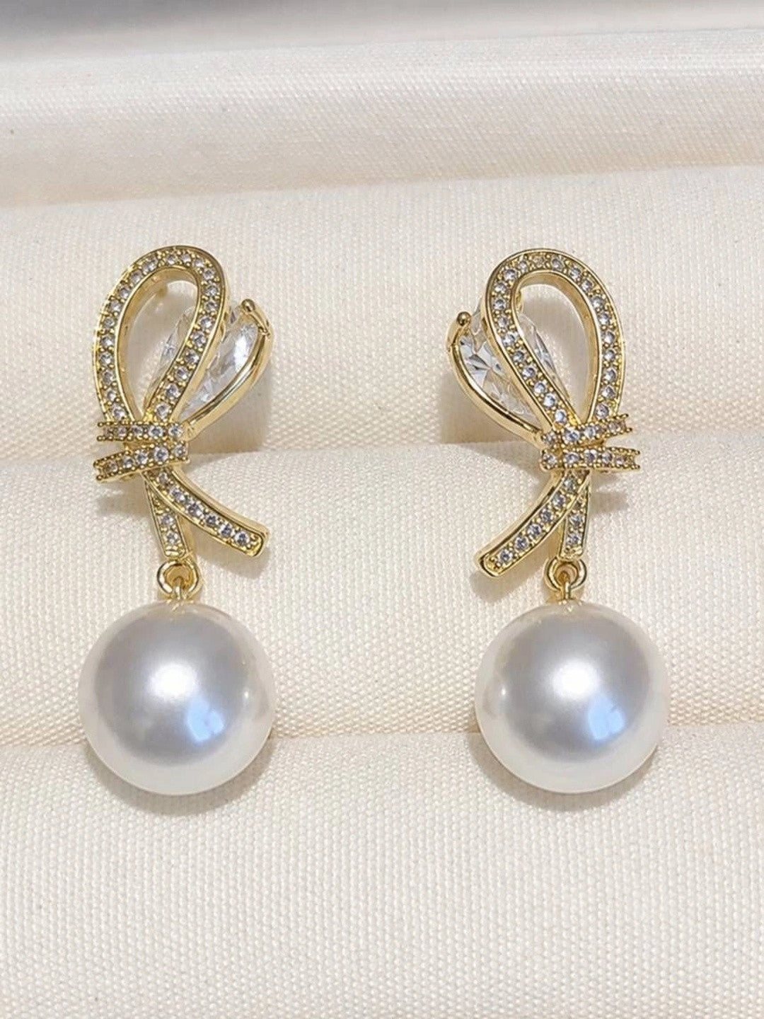 Two gold CZ bow pearl drop earrings laid flat on white fabric showing marquise crystal detail