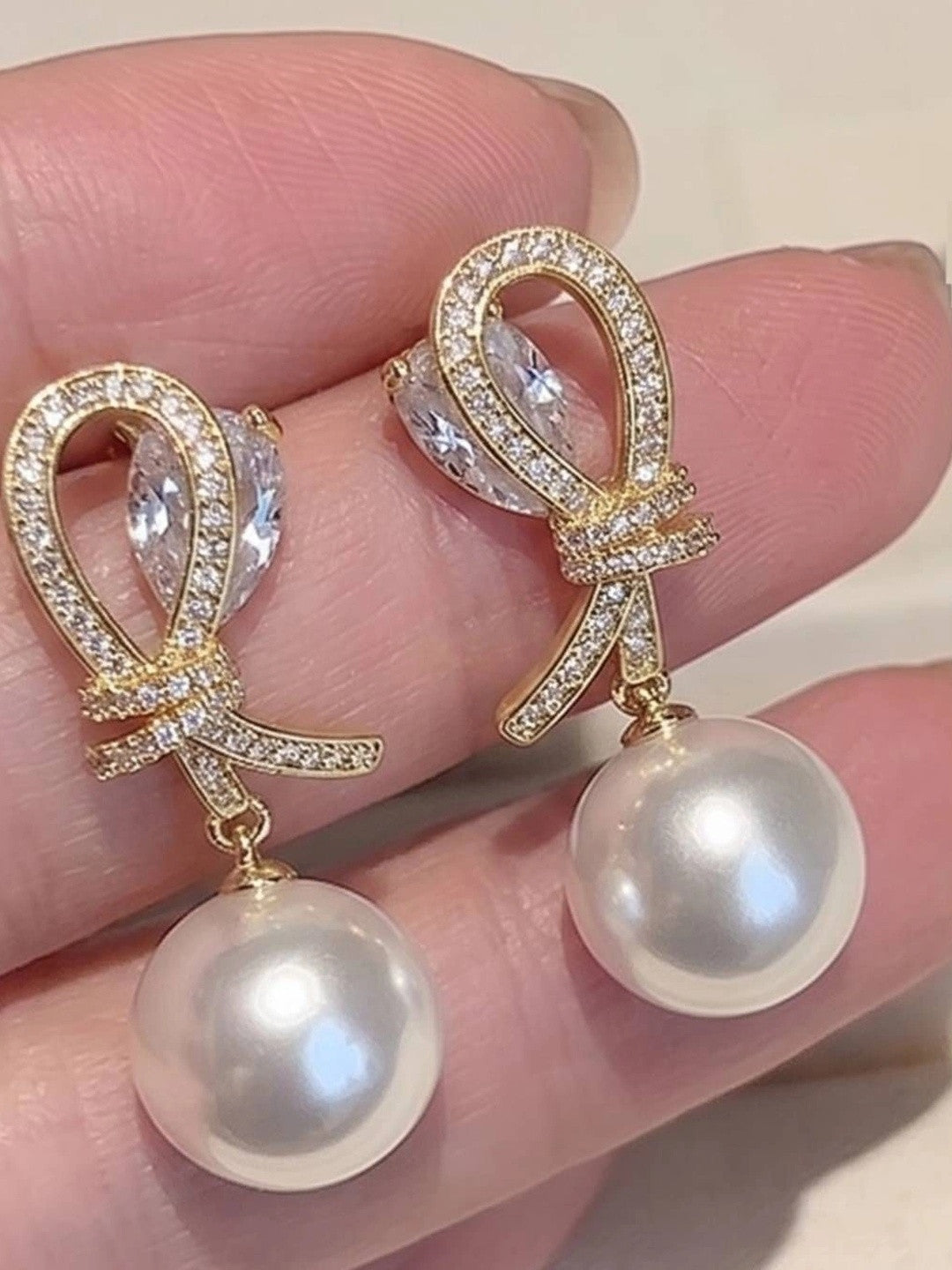 Close-up of gold CZ bow pearl drop earrings held between fingers showing pavé stone detail