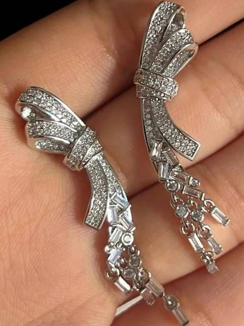 Two silver bow earrings held in hand showing baguette CZ dangles and round stone drops