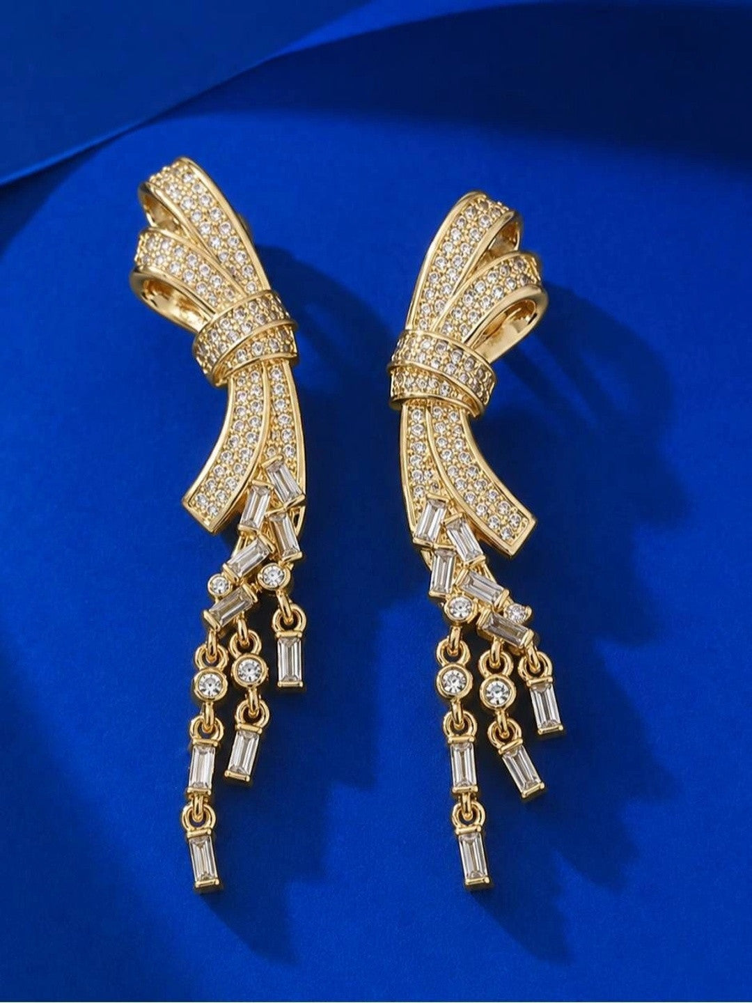 Front view of both gold bow earrings with cascading baguette CZ fringe against a blue satin background