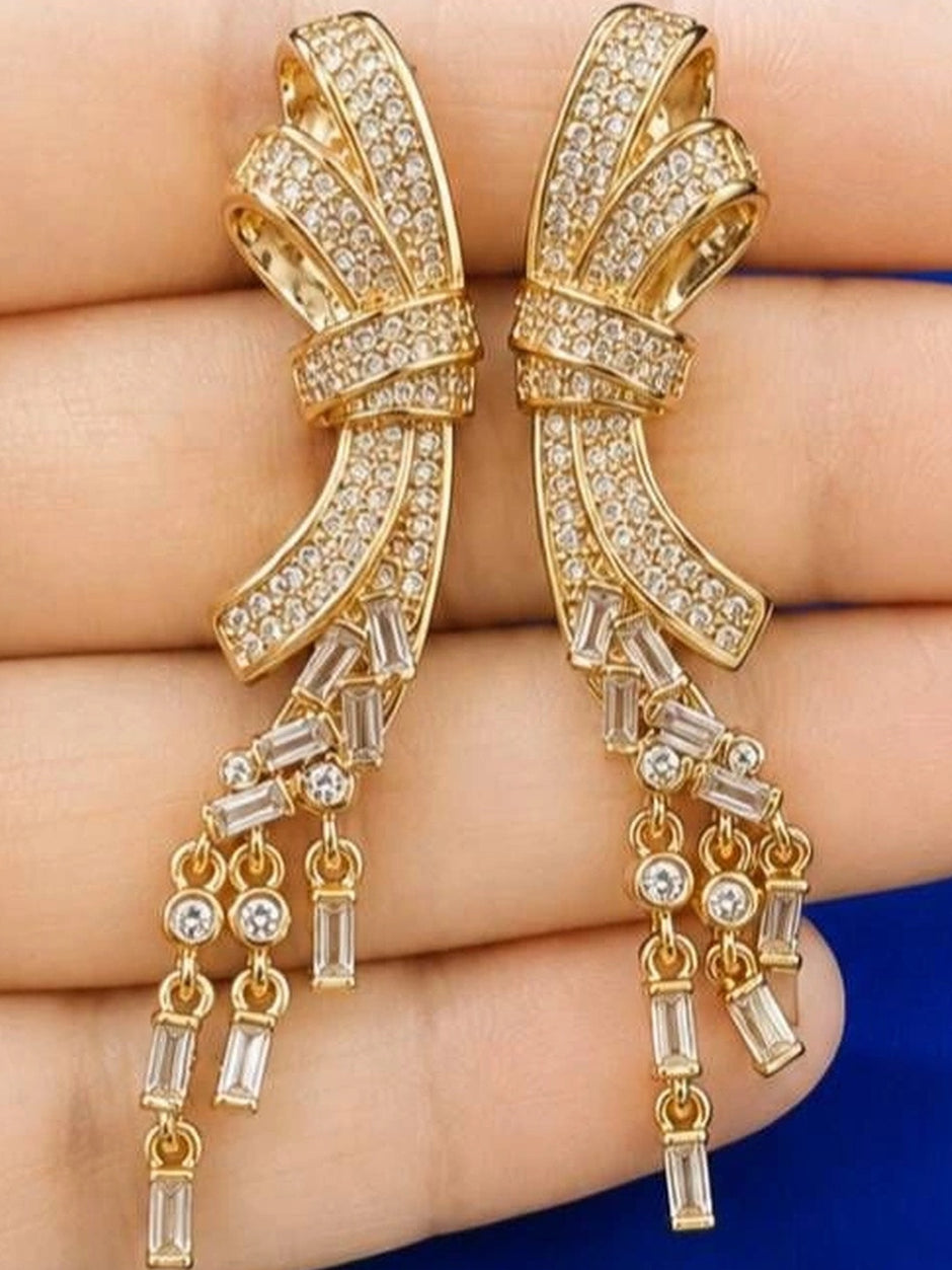 Close-up of gold-plated bow earrings held in a hand showing pavé CZ detail and layered baguette tassels