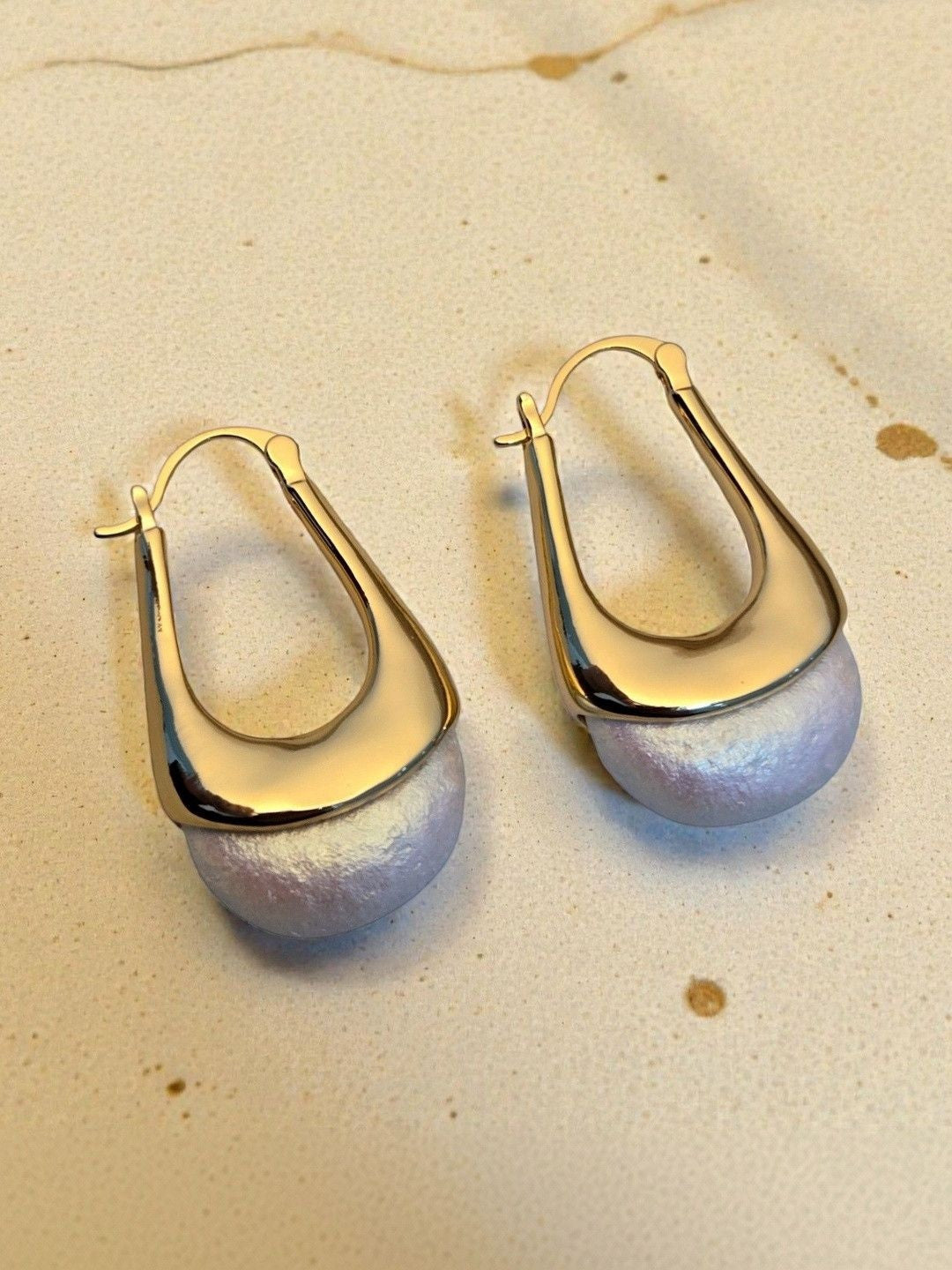 Close-up of gold hoop pearl drop earrings laid flat on a pale textured surface