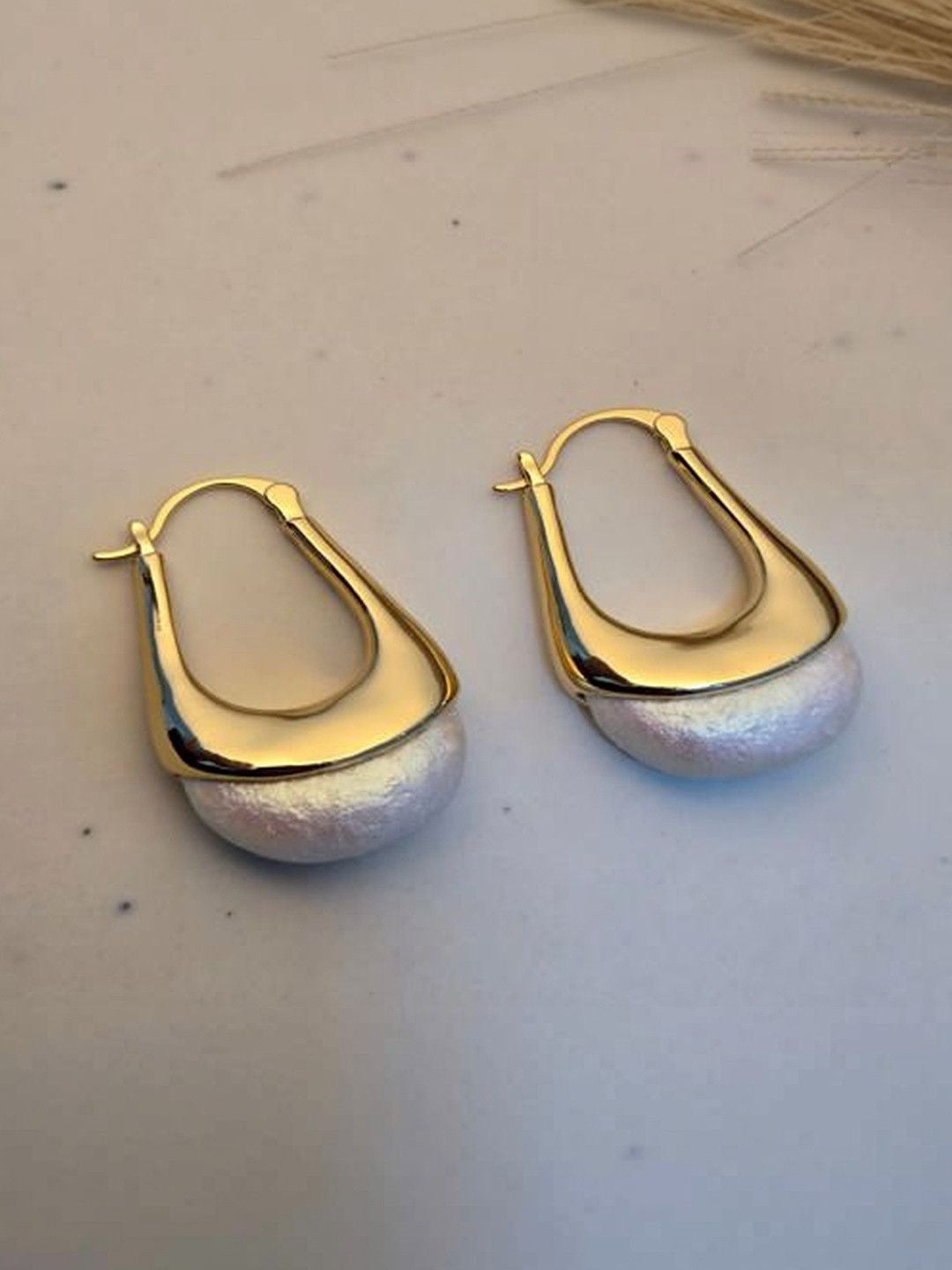Pair of gold and pearl hoop earrings side by side on a white surface near dried grass