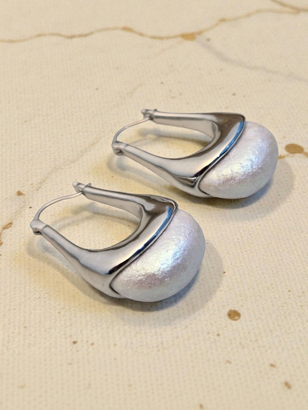 Two silver hoop earrings with iridescent pearl dome base resting on a textured cream fabric background