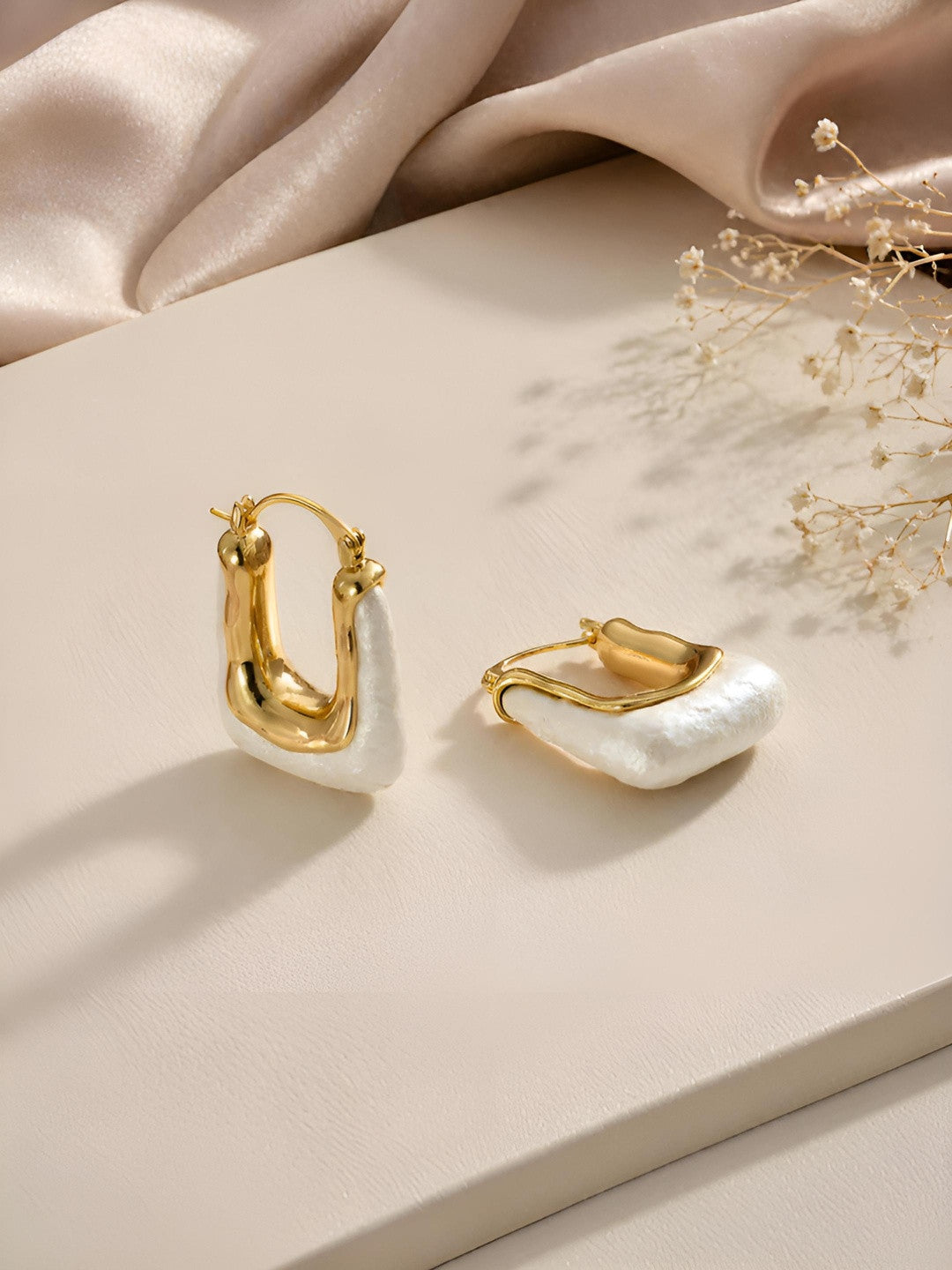 Pair of gold-plated baroque pearl hoop earrings displayed on a cream surface with soft satin and dried flowers in the background