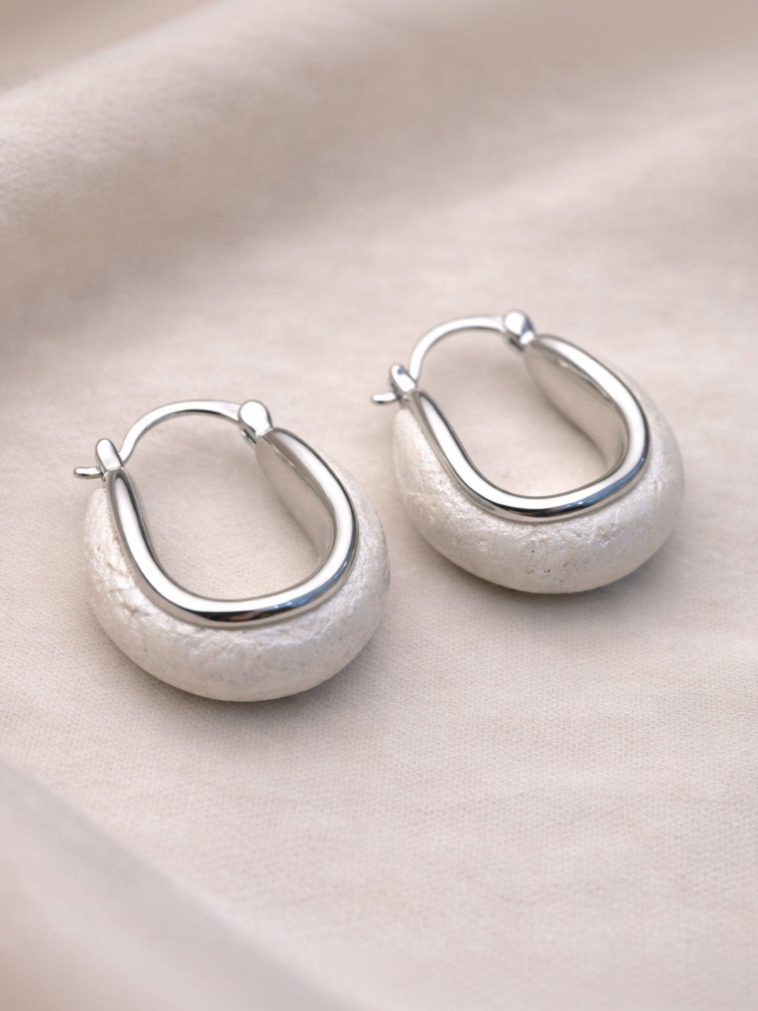 Pair of silver oval hoop earrings with matte stone base resting on a smooth pebble against cream fabric
