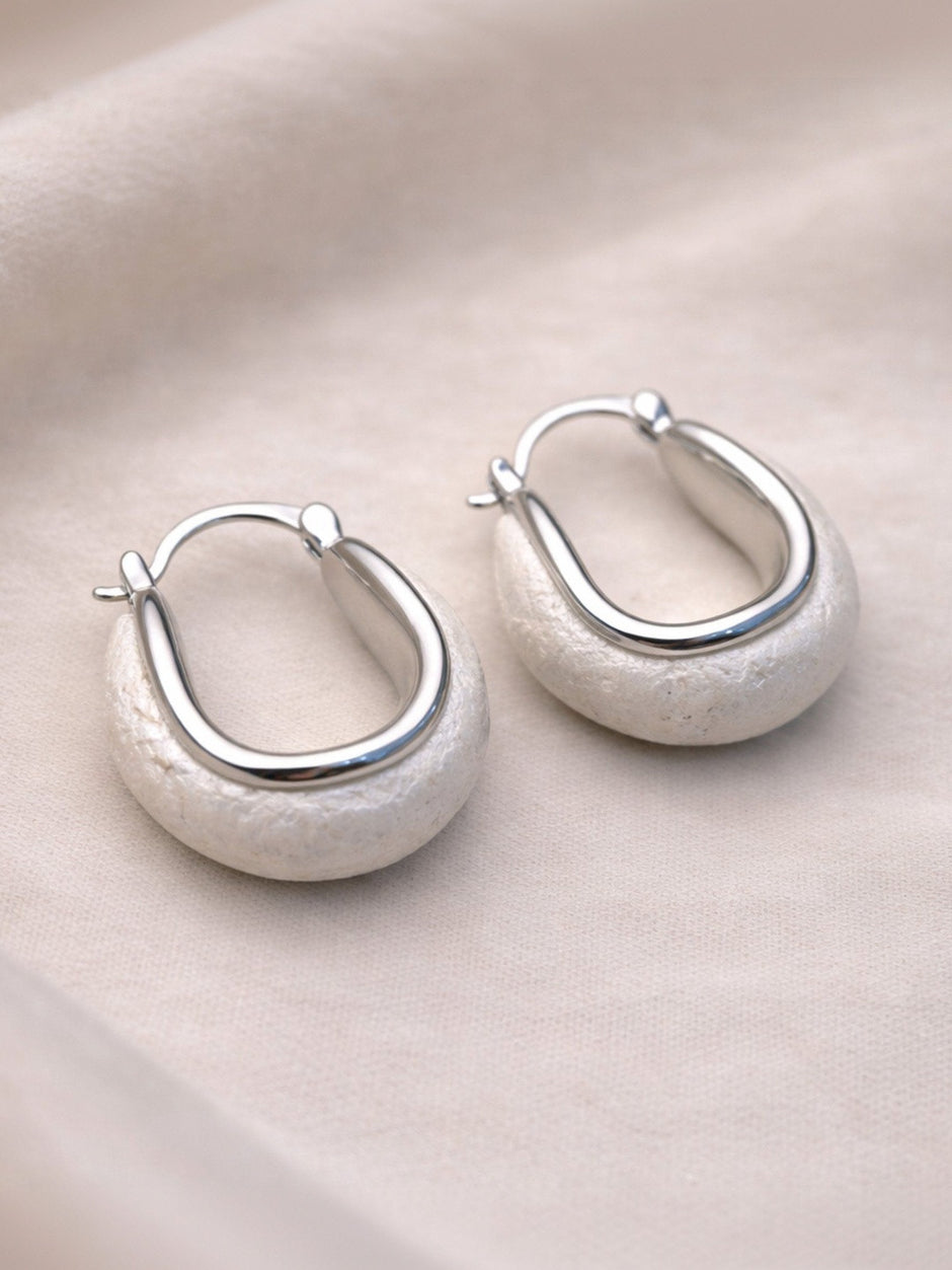 Pair of silver oval hoop earrings with matte stone base resting on a smooth pebble against cream fabric