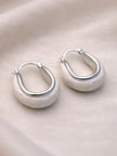 Pair of silver oval hoop earrings with matte stone base resting on a smooth pebble against cream fabric