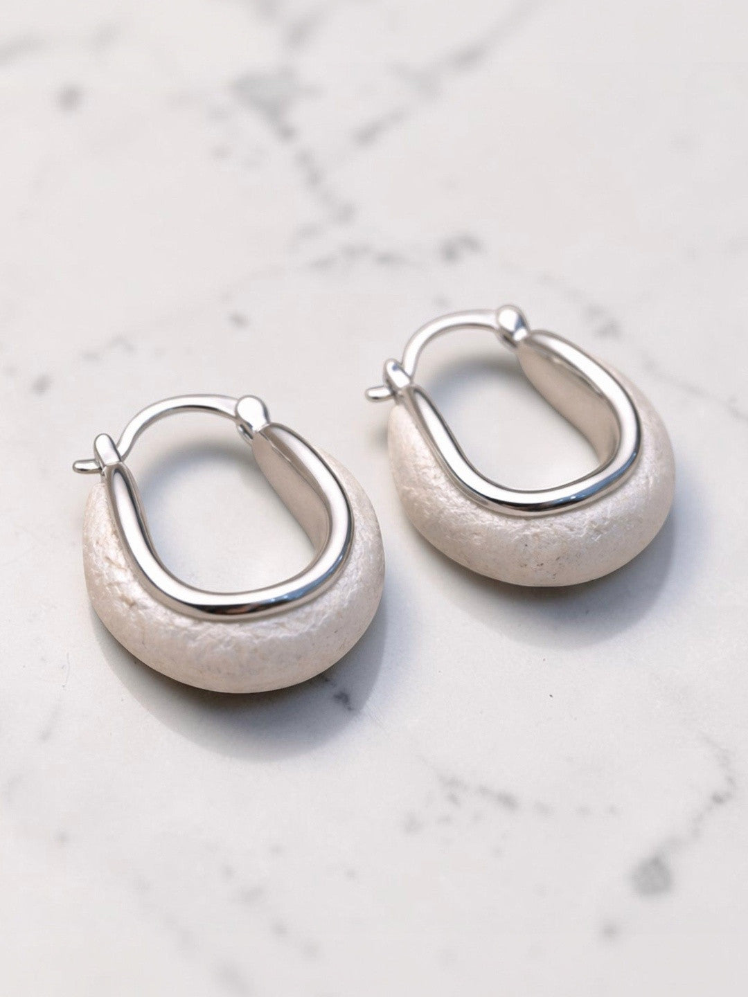 Two silver-tone oval hoop earrings with chunky stone-look base displayed on white marble surface
