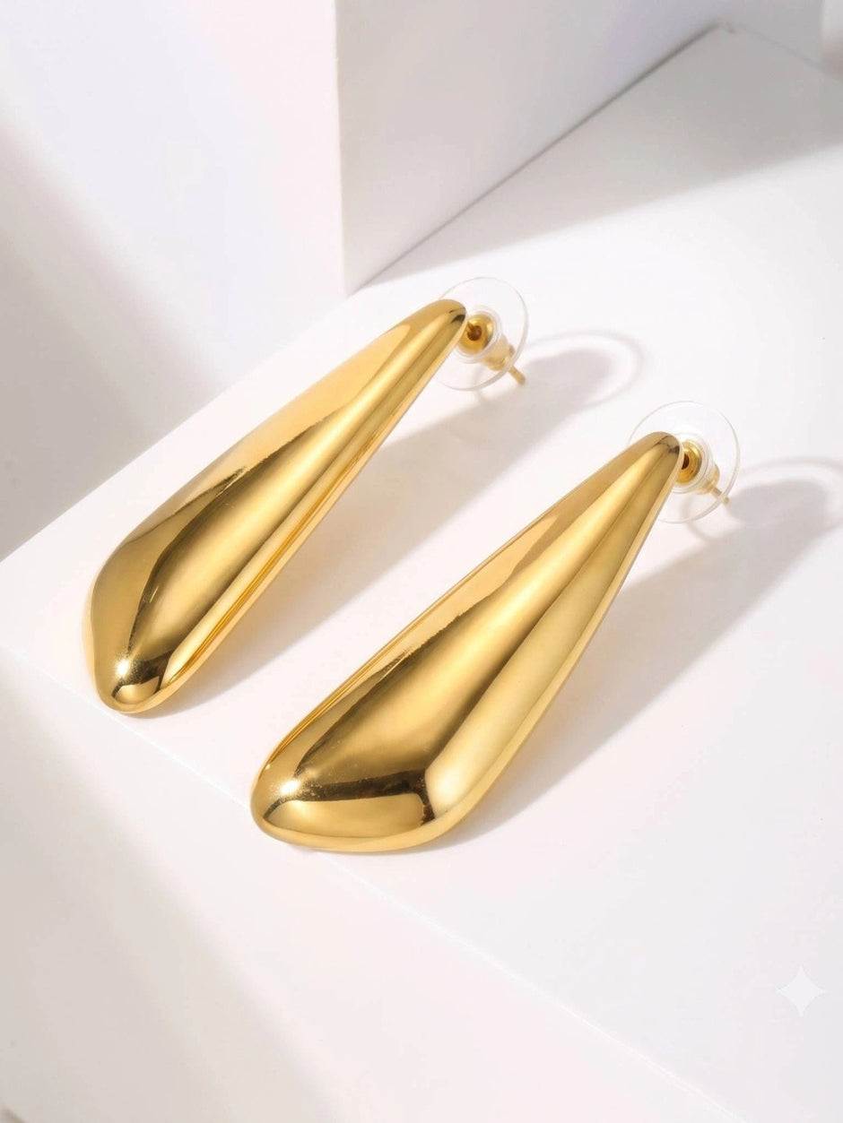 Two elongated gold-toned teardrop stud earrings with push-back stoppers resting on a white block