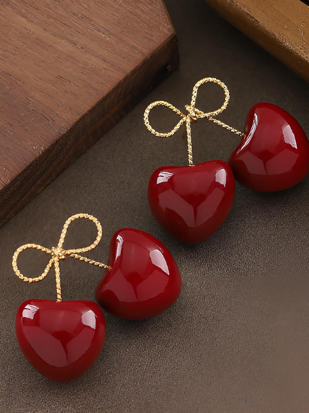 Two pairs of red enamel cherry double-drop earrings with gold-toned bow tops laid on a dark surface beside a wooden block
