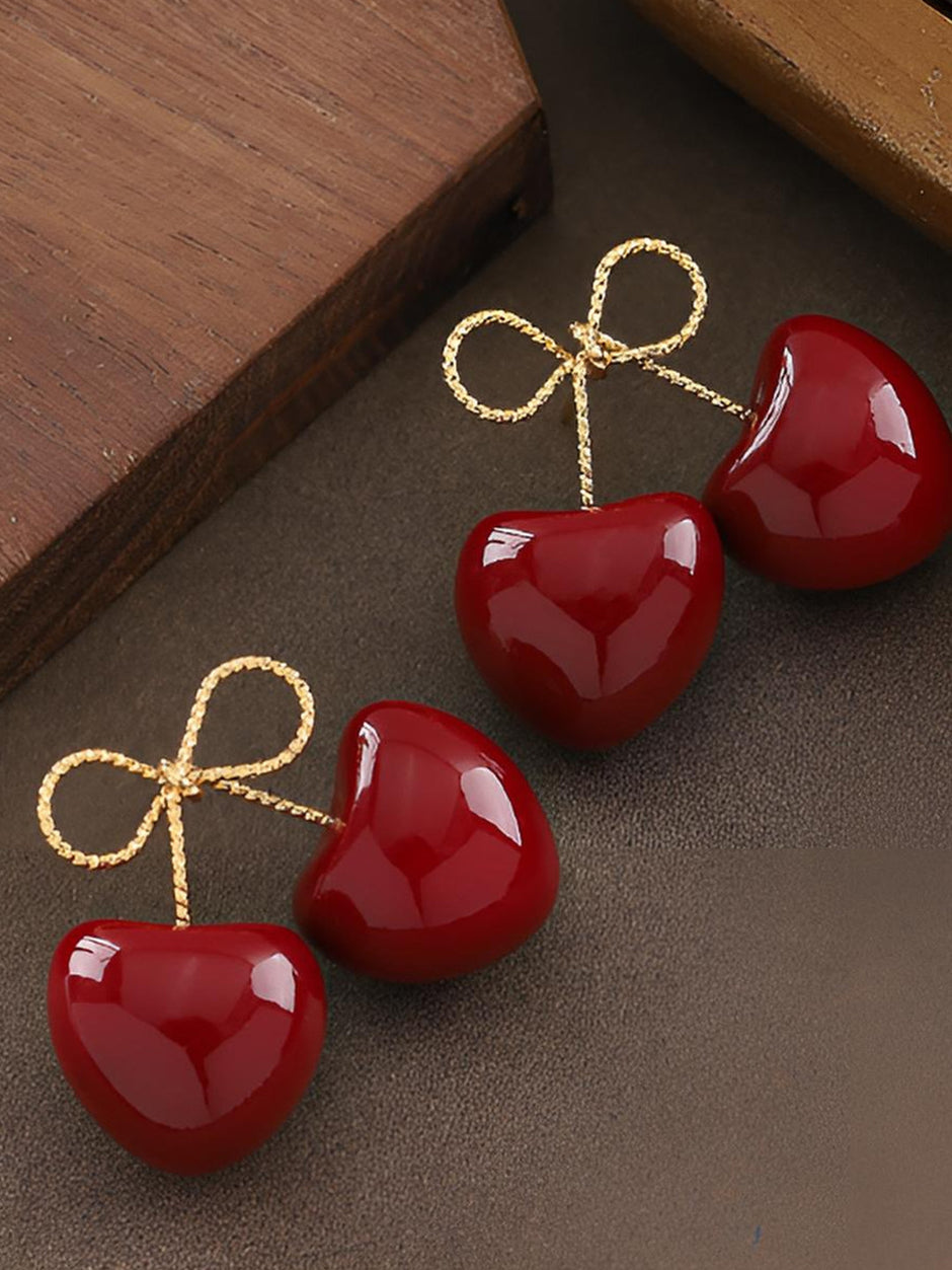 Two pairs of red enamel cherry double-drop earrings with gold-toned bow tops laid on a dark surface beside a wooden block