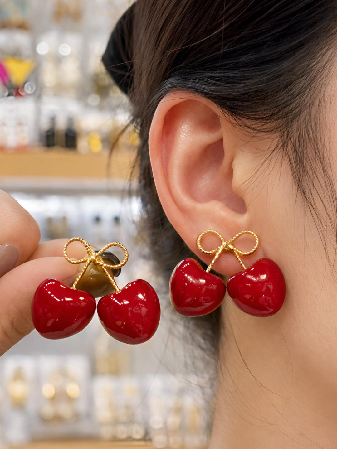 Red enamel cherry bow stud earring worn on a woman's ear with a second earring held up for comparison in a jewellery store