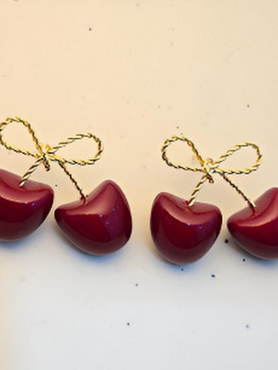 Four red enamel cherry earrings with gold and black twisted-wire bows arranged in a row on a white surface