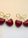 Four red enamel cherry earrings with gold and black twisted-wire bows arranged in a row on a white surface