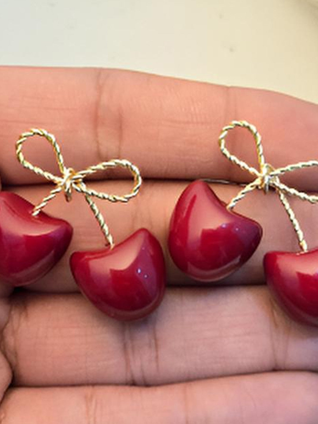 Two pairs of deep red enamel cherry bow earrings resting on an open palm showing scale and finish detail