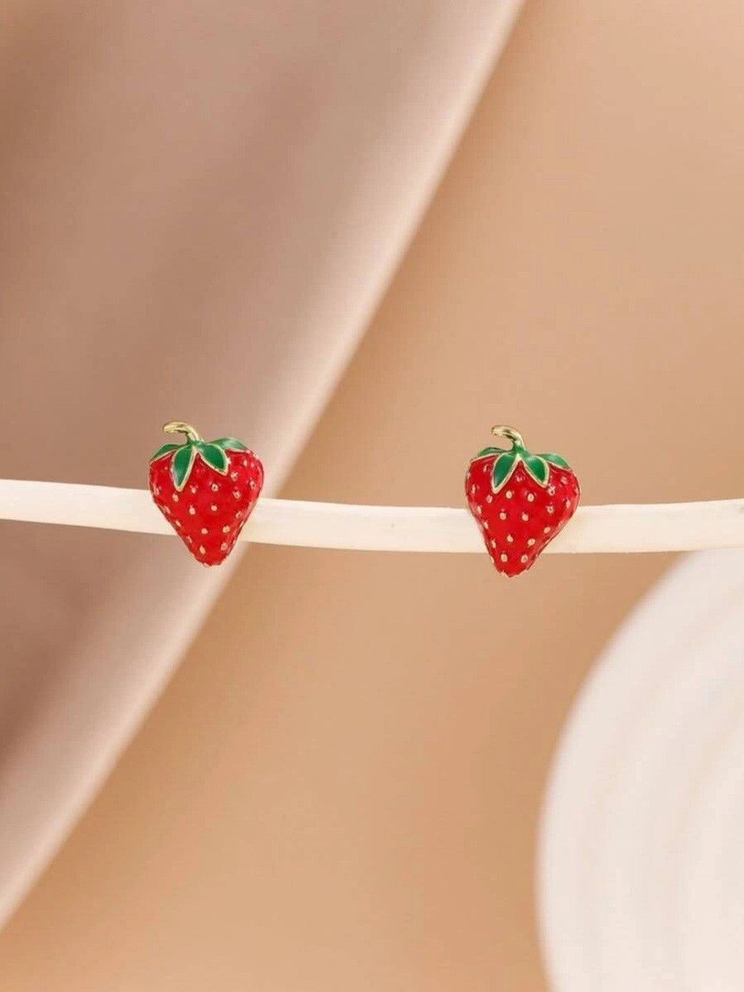 Pair of red enamel strawberry stud earrings resting on a cream surface against a soft pink background