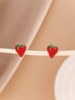 Pair of red enamel strawberry stud earrings resting on a cream surface against a soft pink background