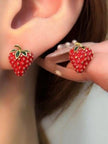 Red enamel strawberry stud earring worn on ear alongside a second stud held between manicured fingers