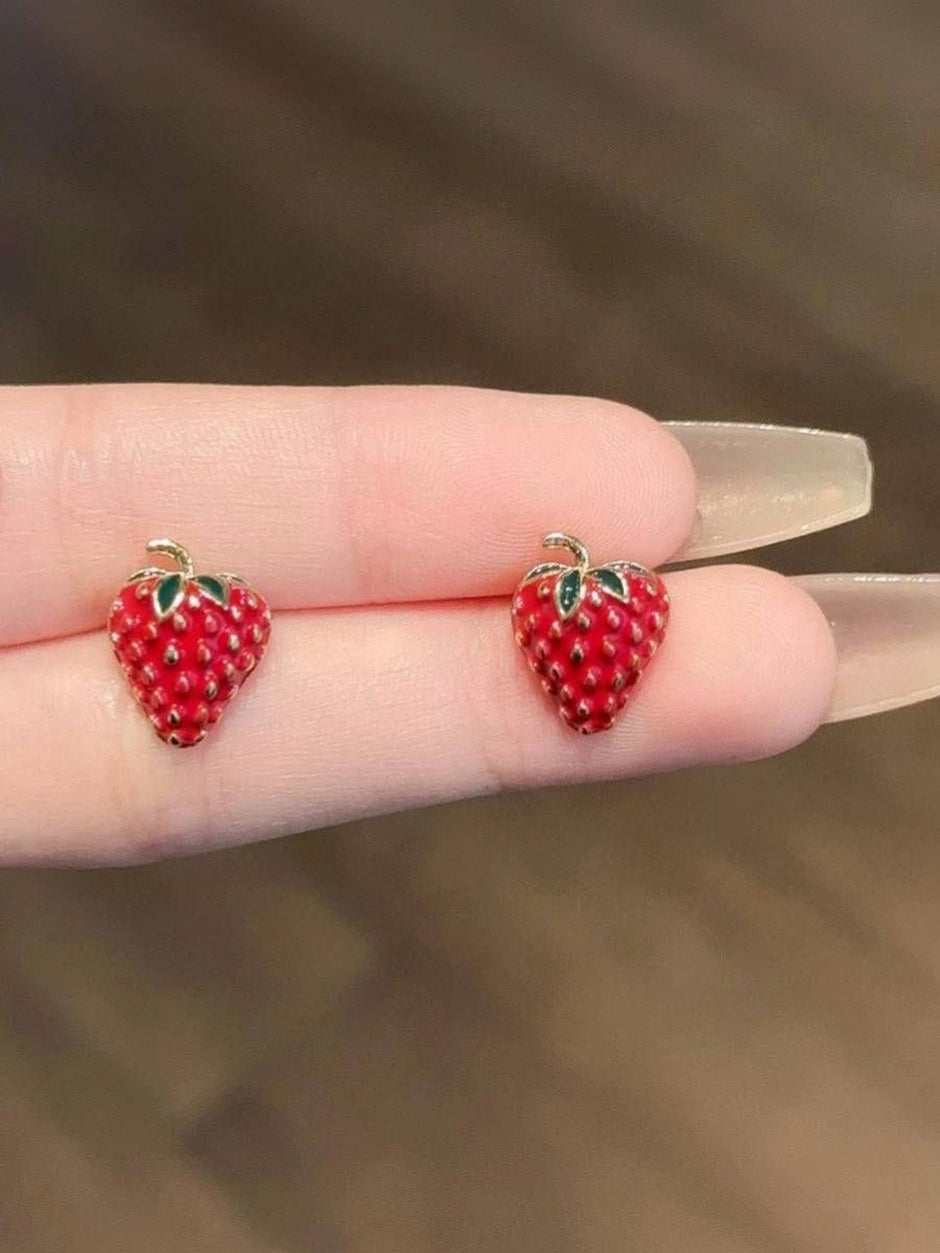 Close-up of two red enamel strawberry stud earrings balanced on fingertips showing raised seed texture detail
