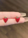 Close-up of two red enamel strawberry stud earrings balanced on fingertips showing raised seed texture detail