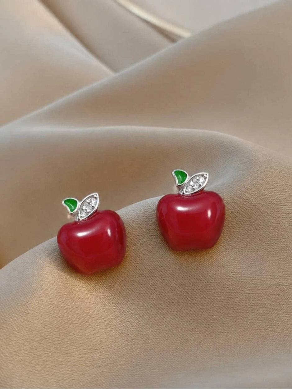 Two red enamel apple studs with green and crystal leaf tops resting on beige fabric