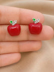 Red enamel apple stud earrings held between fingertips showing size and glossy finish