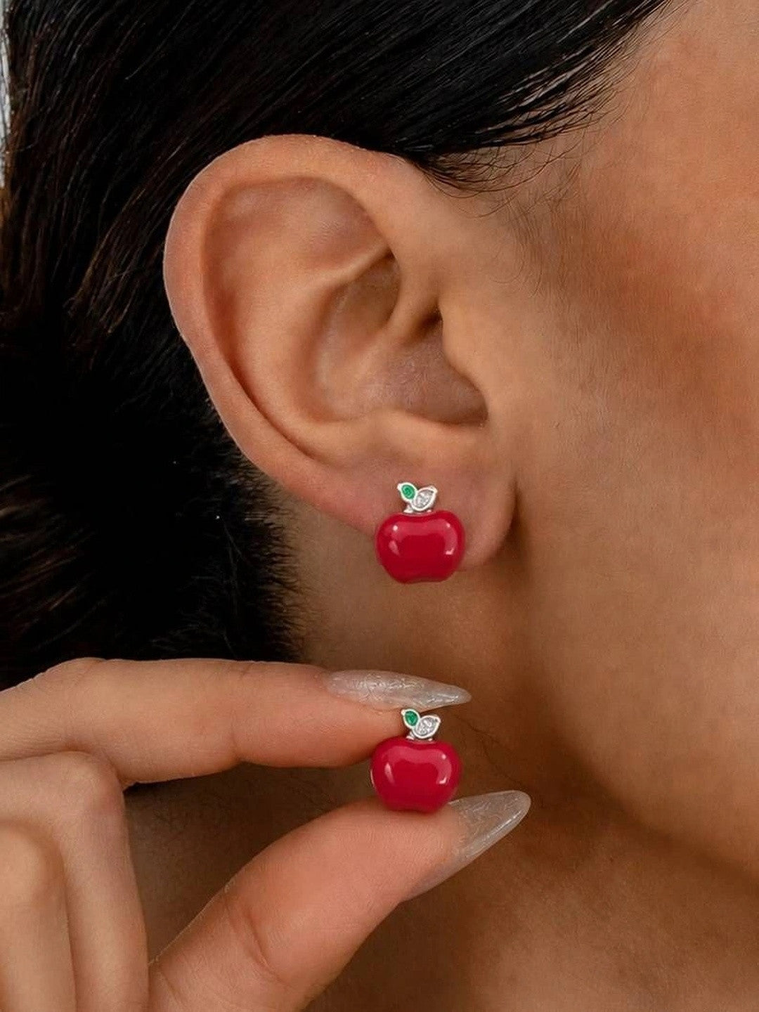 Red apple stud earring worn on ear with matching stud held between fingers showing wearable size