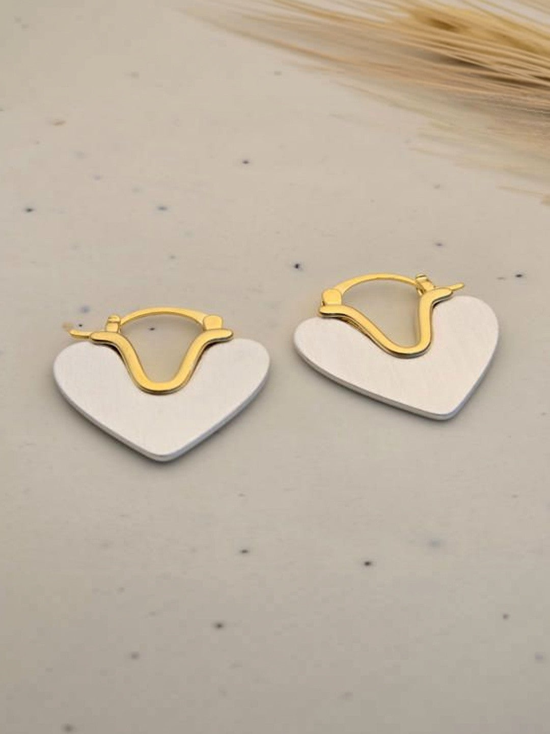 Pair of two-tone heart hoop earrings with gold-tone lever-back and brushed silver heart charm on speckled beige surface
