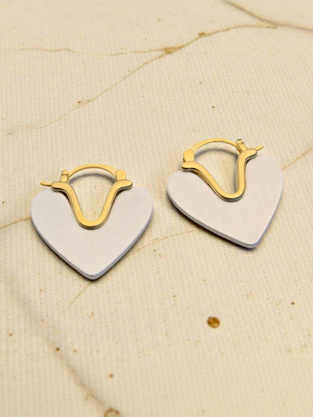 Two-tone heart hoop earrings laid flat on cream fabric showing gold hoop detail and silver heart front