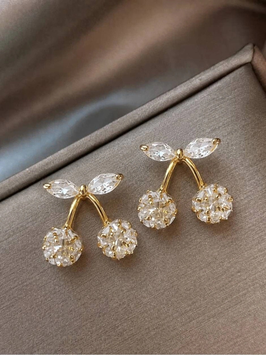 Pair of gold-plated crystal cherry stud earrings laid flat on taupe satin fabric showing both pieces side by side