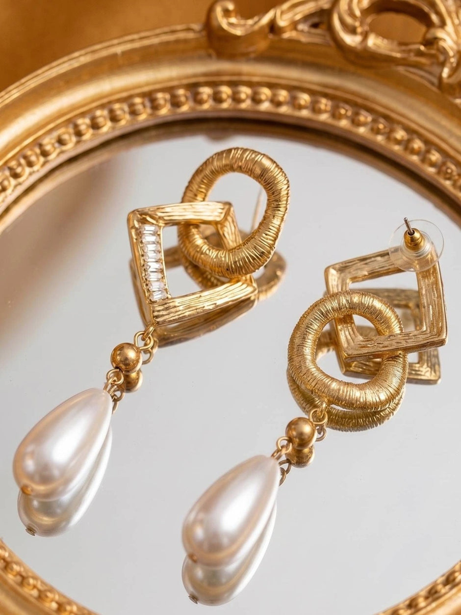 Pair of gold-plated geometric drop earrings with faux pearl teardrops lying on a round mirror