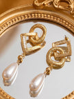 Pair of gold-plated geometric drop earrings with faux pearl teardrops lying on a round mirror