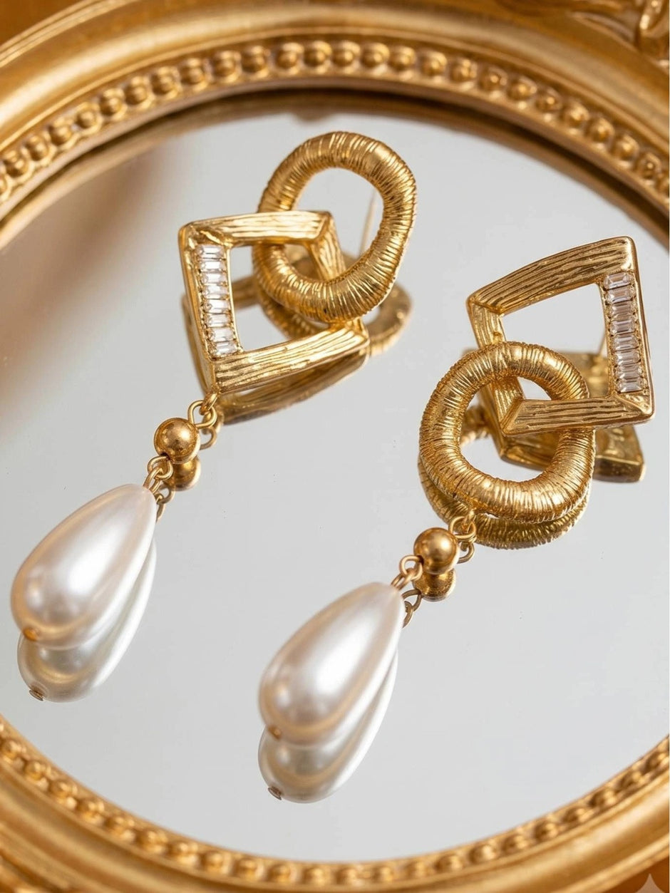 Two gold textured geometric earrings with pearl drops resting on an ornate round mirror