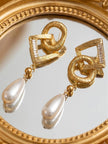Two gold textured geometric earrings with pearl drops resting on an ornate round mirror