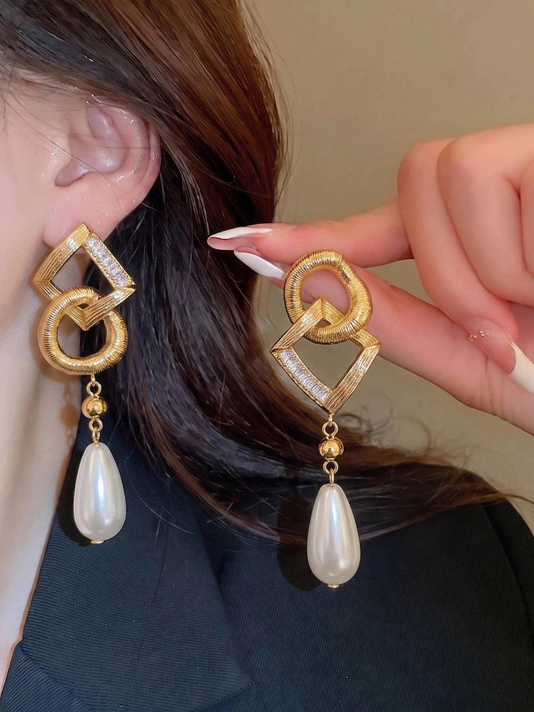 Woman wearing gold geometric pearl drop earring on ear, holding second earring beside it