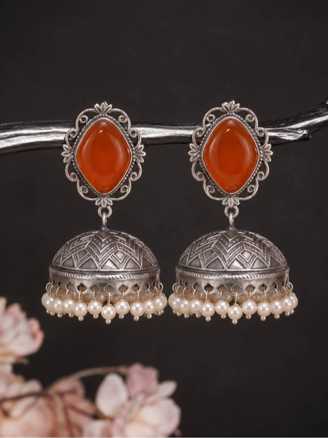 Pair of oxidised silver jhumka earrings with orange carnelian stone tops and ivory pearl fringe, displayed on a dark branch