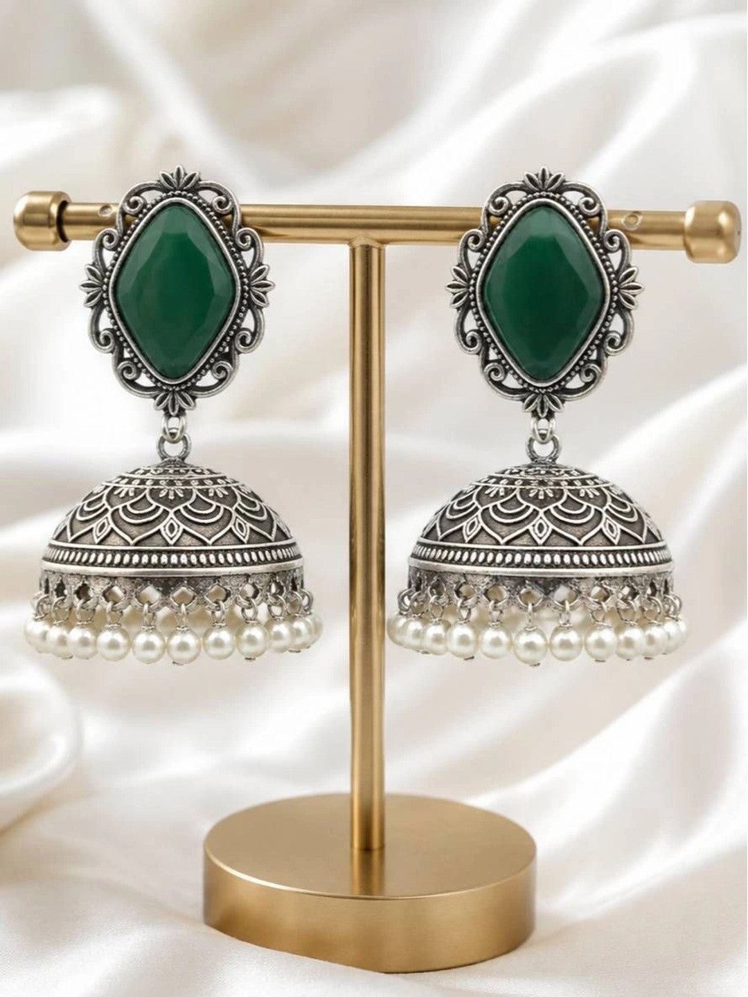 Pair of oxidised silver jhumka earrings with green stone tops and white pearl drops displayed on a gold earring stand